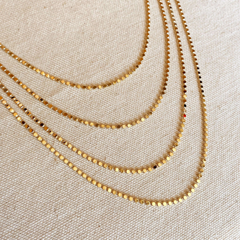 angled view of multiple layered tarnish free flat ball 18 karat gold filled necklaces laying flat on a canvas background showing brilliance and shiny gold metal