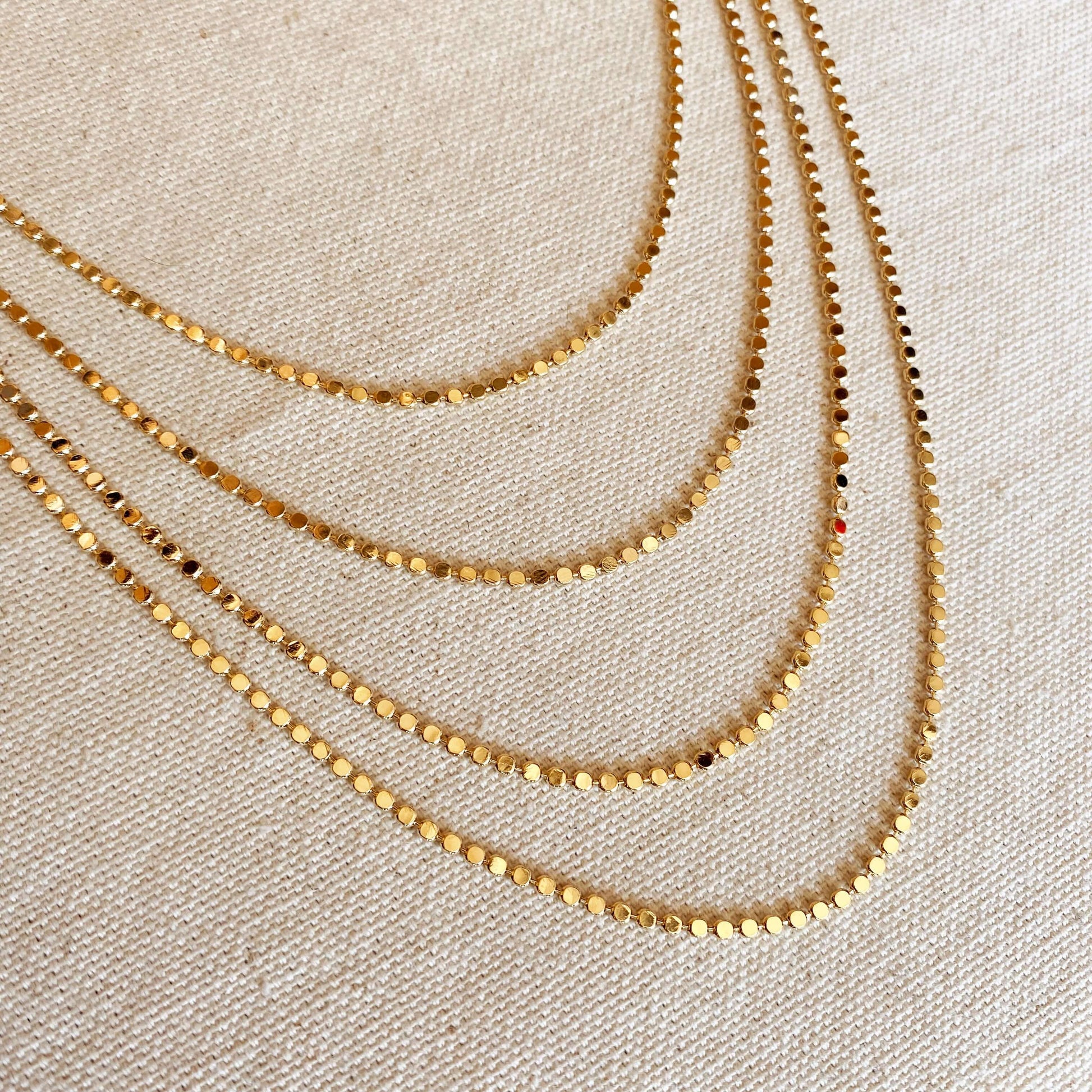 angled view of multiple layered tarnish free flat ball 18 karat gold filled necklaces laying flat on a canvas background showing brilliance and shiny gold metal